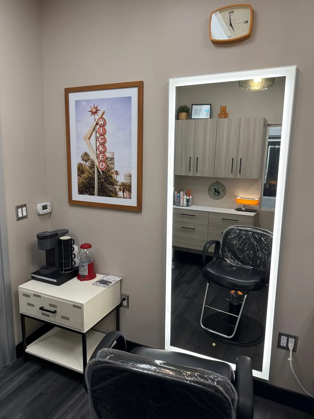 Styling chair facing a lighted floor mirror next to a framed Wicked sign print and a coffee station